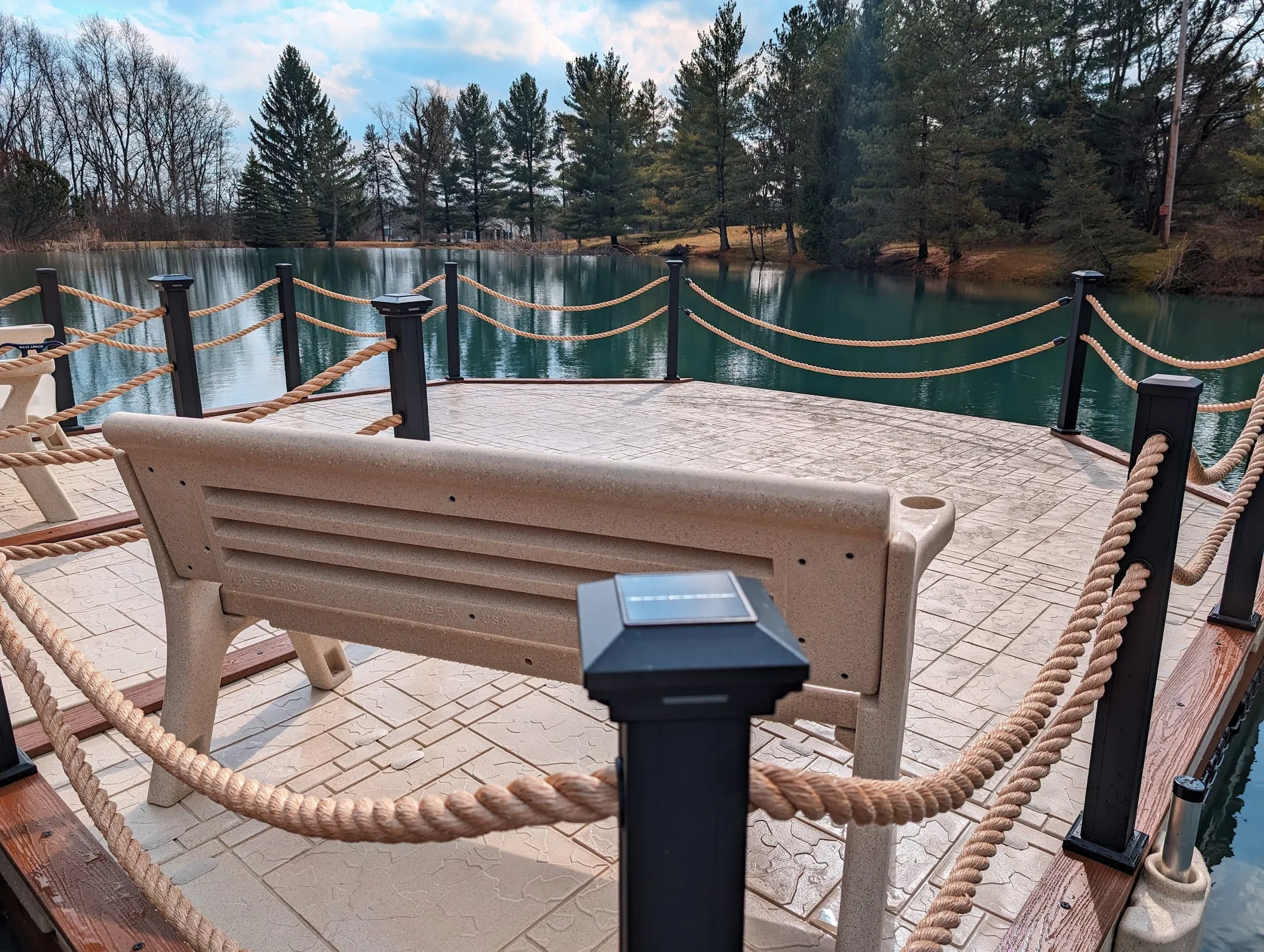 AquaLife custom Wave Armor dock — pond view and seating Wide view across a beige bench and textured AquaLife Wave Armor dock toward a wooded pond, with tan rope detailing and dark posts